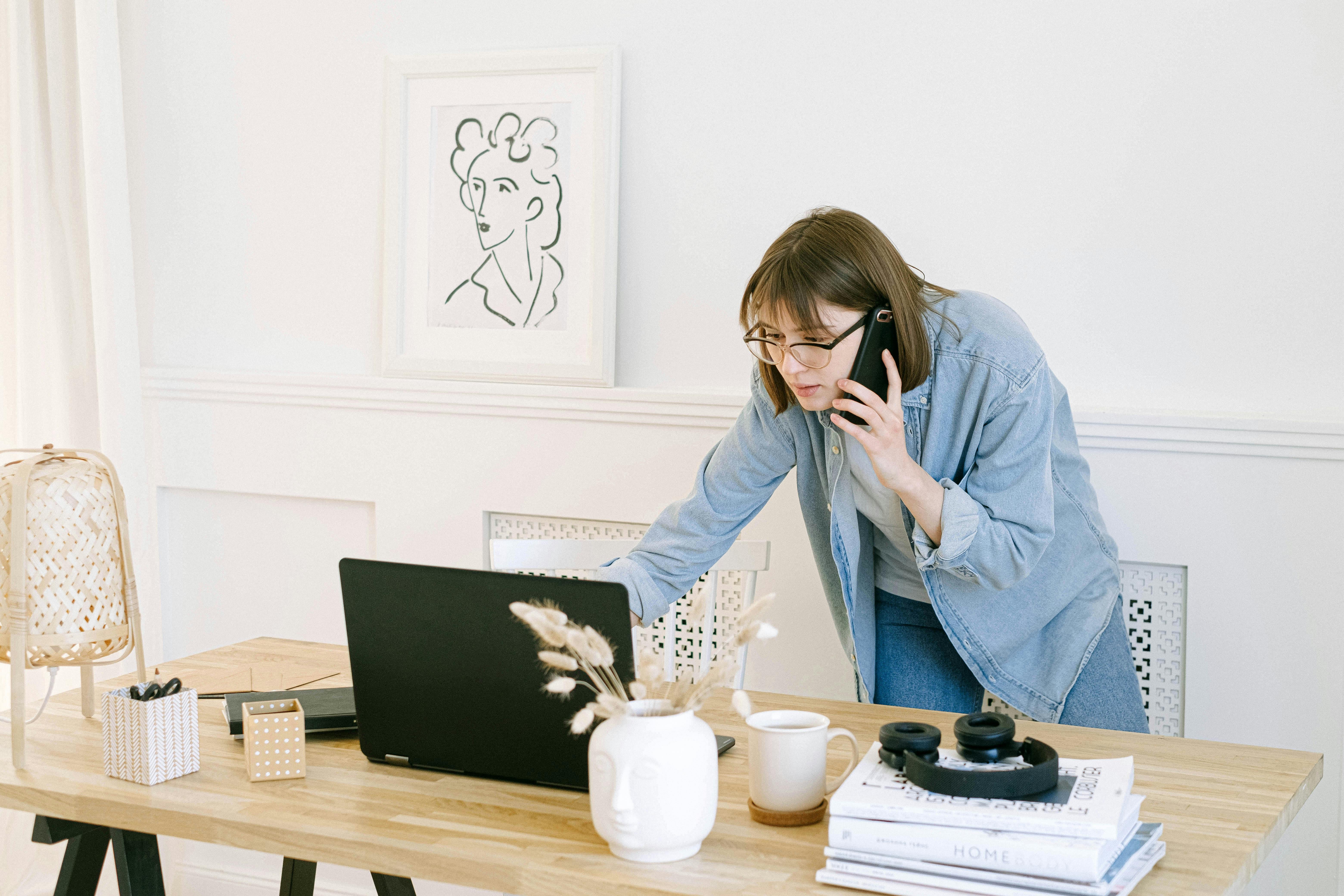 Woman in light blue denim shirt and glasses leaning over wooden desk speaking on phone while viewing laptop, in bright minimalist home office with white walls, framed portrait art, and desk accessories including white mug with pampas grass, stacked books, and wireless headphones.