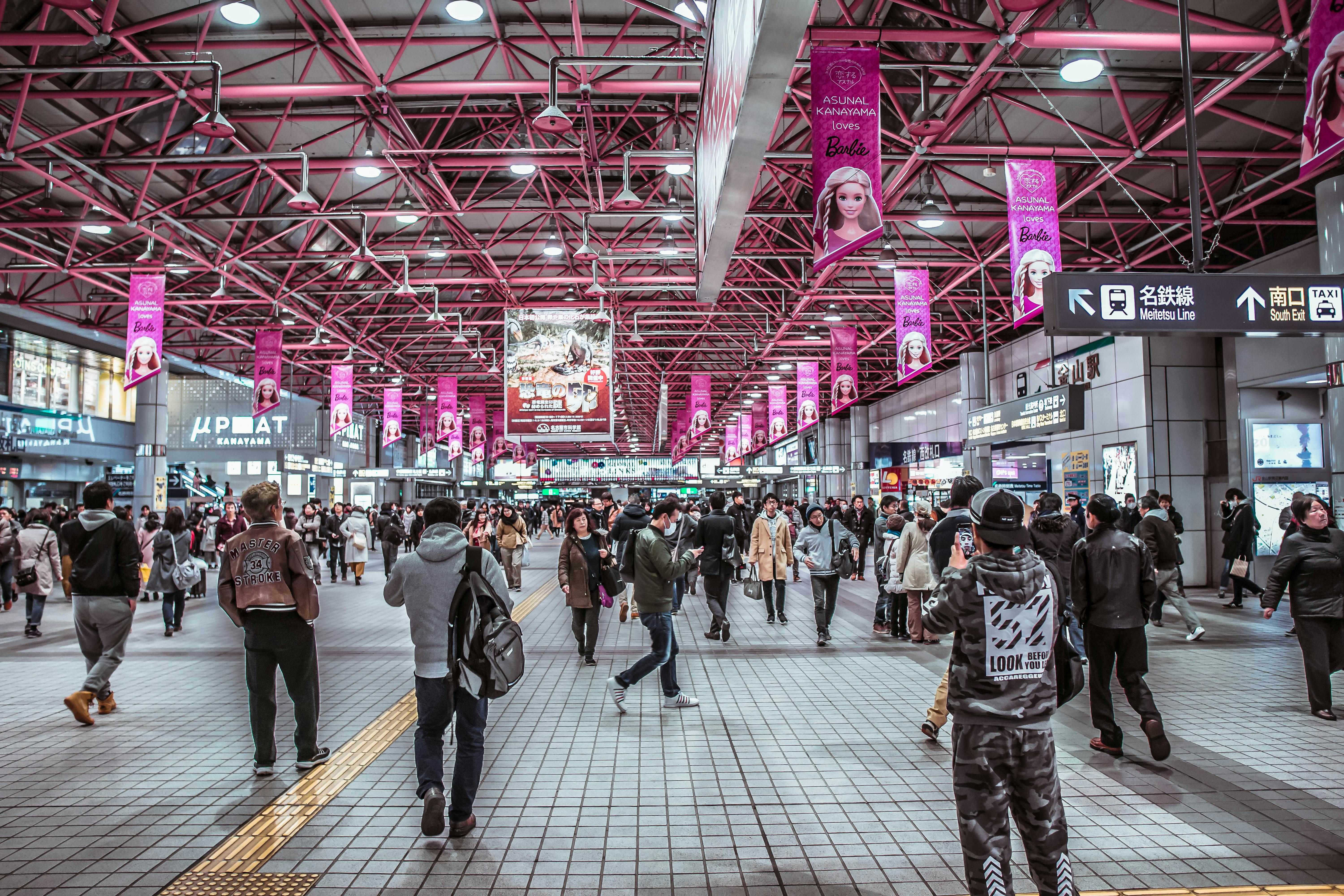 Busy Japanese train station concourse with young diverse crowd walking through modern pink-lit retail space, Barbie campaign banners hanging from industrial ceiling, shops visible on sides